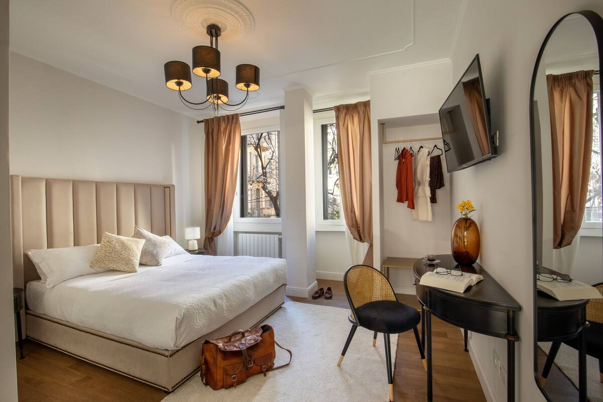 A bedroom with a large bed, a desk with a chair, a mirror, and a view of the outdoors.