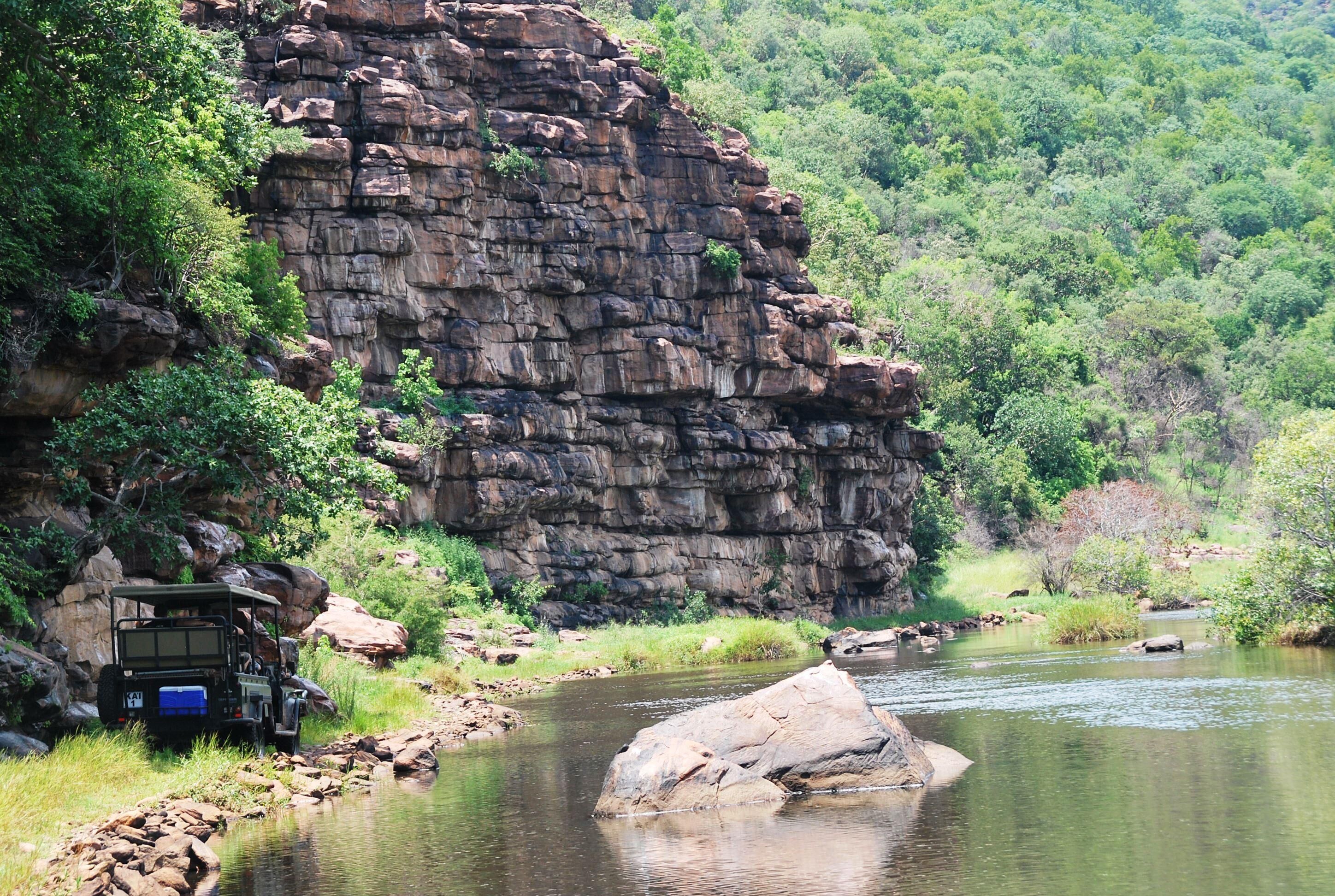 A jeep parked by a river with a rocky cliff in the background.