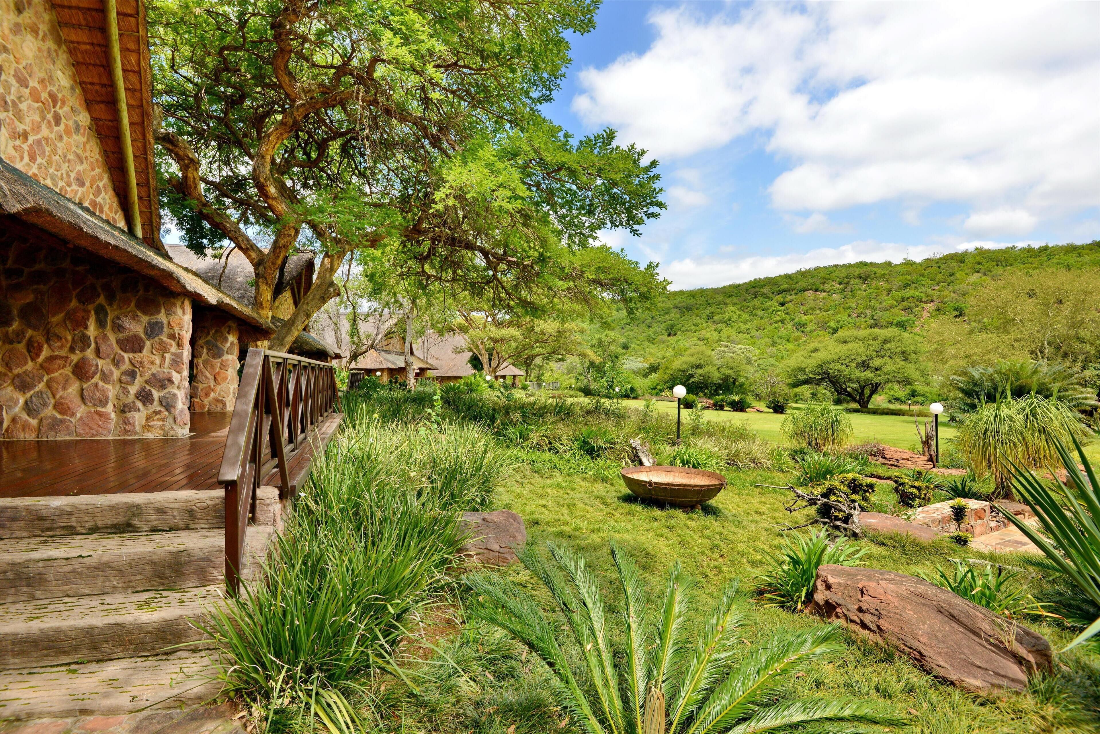 A stone building with a wooden staircase leading to a deck, surrounded by lush greenery and a hilly landscape.