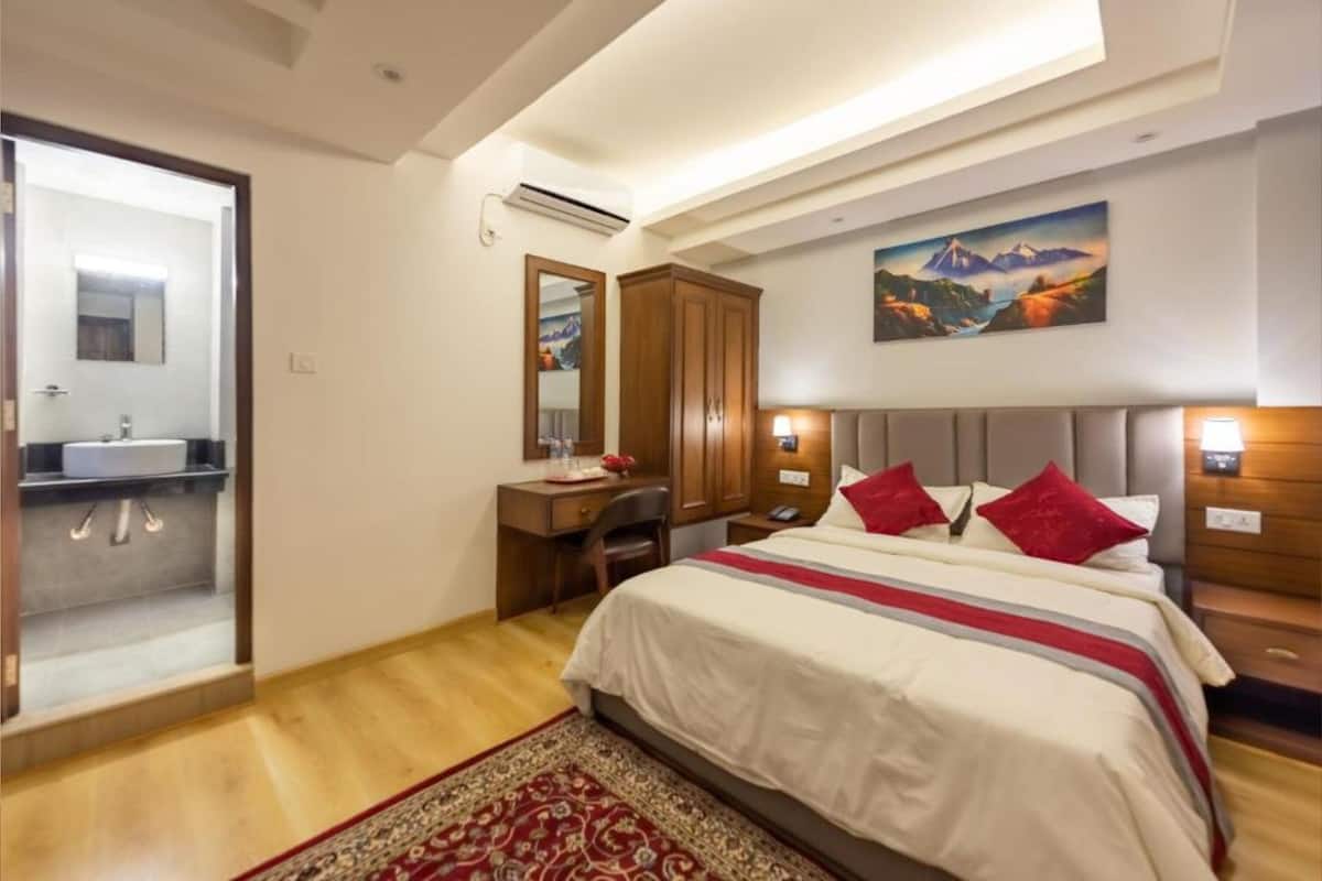 A hotel room with a bed, a desk, a chair, a mirror, a painting, and a bathroom visible through an open door.