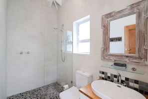 Casa de banho com cabine de duche em vidro, sanita, lavatório com prateleira em vidro e espelho com moldura rústica.