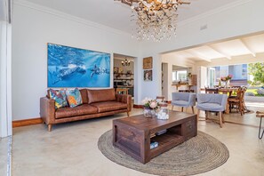 A spacious living area with a large wall-mounted artwork, a brown leather sofa, a wooden coffee table, and a dining area with a table and chairs.