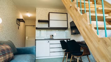 Un espai habitable compacte amb zona de cuina americana, sofà, menjador i escala de fusta.