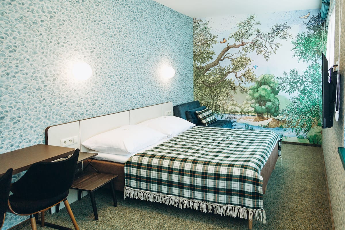 Une chambre à coucher avec un lit, un bureau et une chaise. Un mur est décoré d’une fresque représentant un paysage naturel.