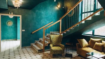A room with teal walls, a patterned armchair, a sofa with cushions, a staircase with wooden railings, and a chandelier.