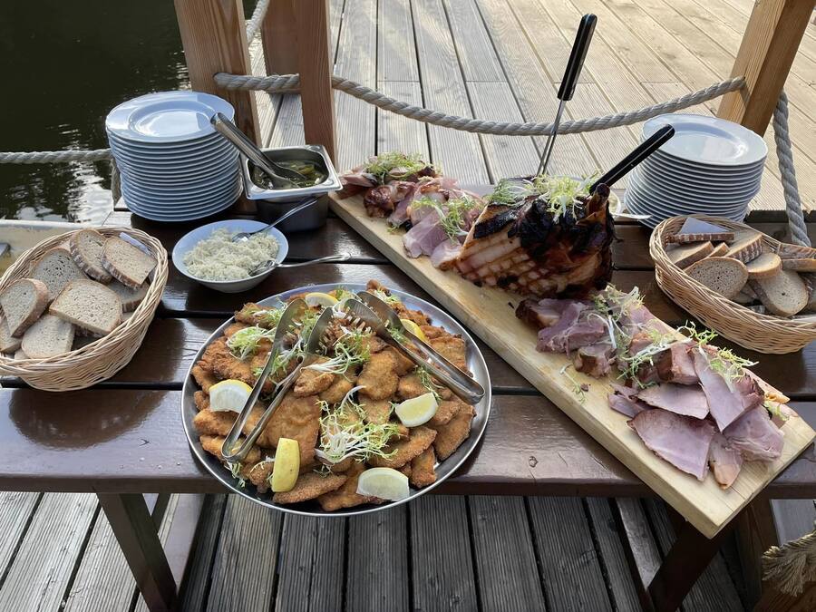 Ein Holztisch mit verschiedenen Speisen, darunter Brot, Fleisch und einer Portion frittiertem Fisch, auf einer Anlegestelle.
