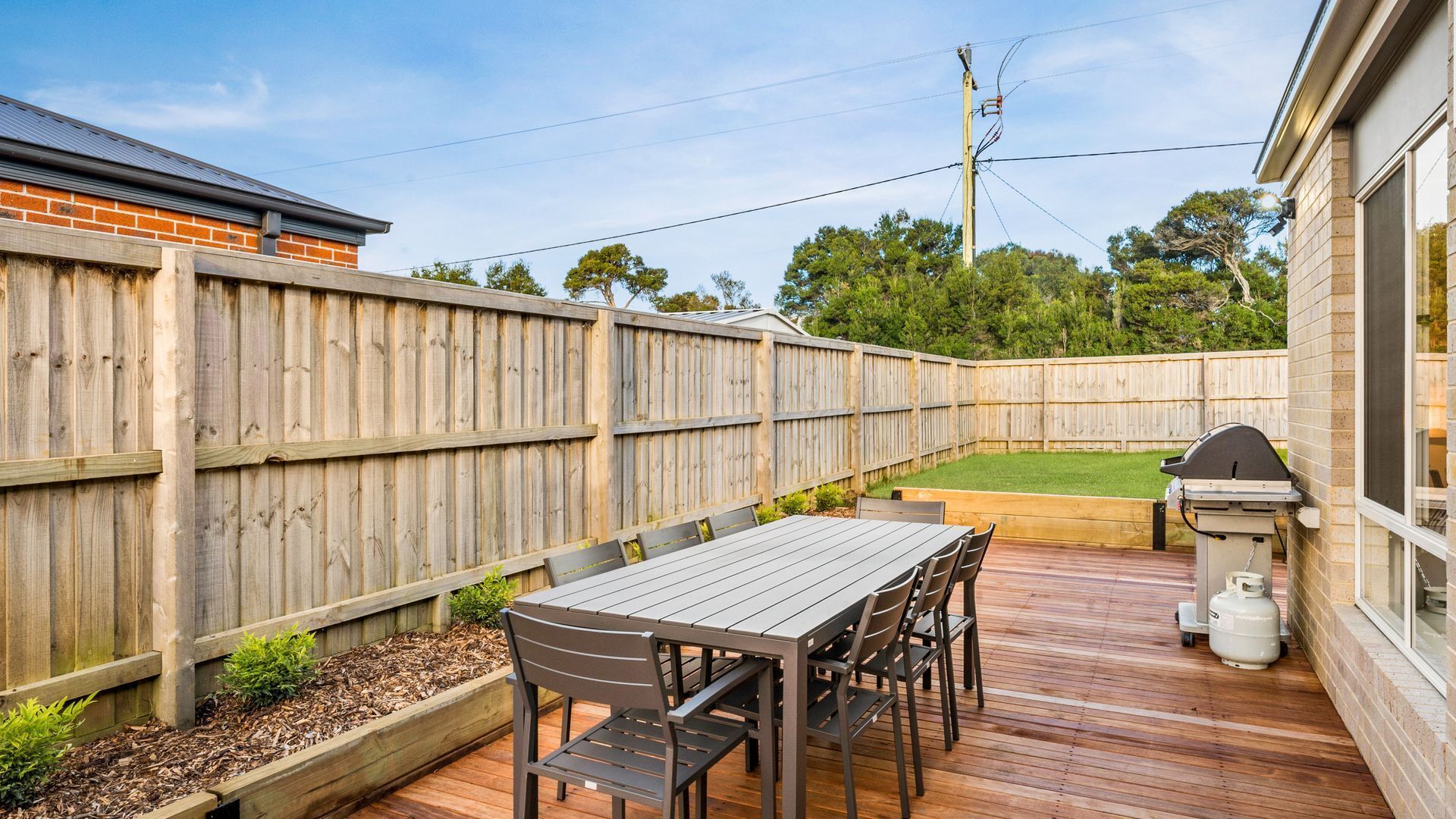 Una terraza de madera con una mesa y sillas, una parrilla de barbacoa y una casa de ladrillo al fondo.