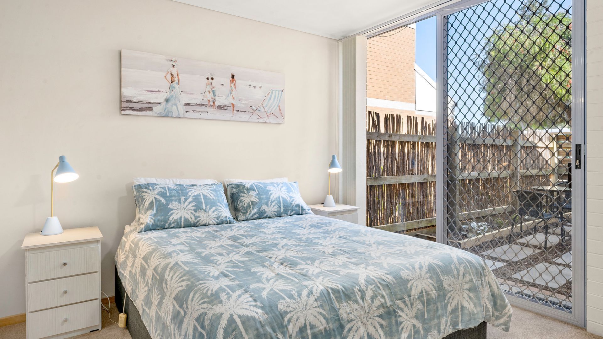 A bedroom with a bed, two bedside tables, a nightstand, a painting, and a view through a glass door to an outdoor area with a fence.