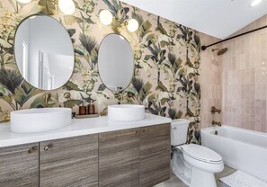 A bathroom with a floral wallpaper, two round mirrors, a white sink, a toilet, and a bathtub. - Magnificent Poolside Views – Private Palm Springs Escape with Spa, Fire Pit & Citrus Garden (Palm Springs)
