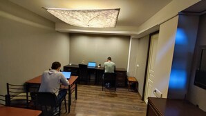 Een kamer met twee bureaus, waarvan er één een laptop en de ander een smartphone heeft. Er is een stoel, een klein tafeltje en een hanglamp.