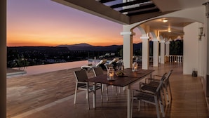 Une terrasse offrant une vue sur le coucher de soleil, une table en bois avec des chaises et un espace piscine.