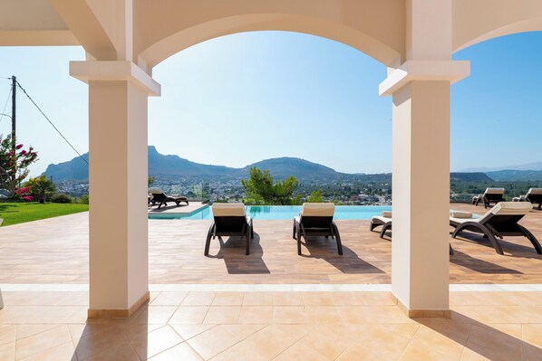 A covered outdoor area with lounge chairs and a pool, overlooking a mountainous landscape.