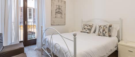 Una camera da letto con un letto bianco, un comodino, una sedia e vista sul balcone esterno.