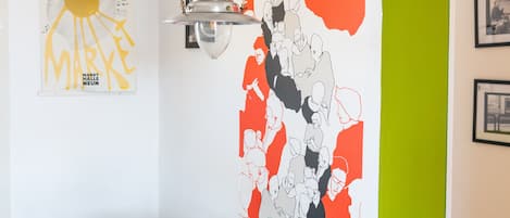 A dining area with a table set for two, a wall with a colorful abstract mural, and a hanging light fixture.
