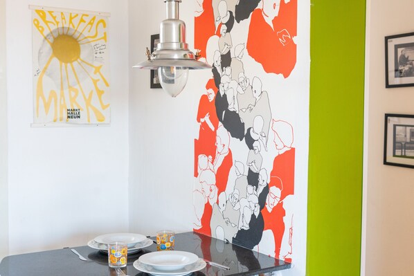 A dining area with a table set for two, a wall with a colorful abstract mural, and a hanging light fixture.