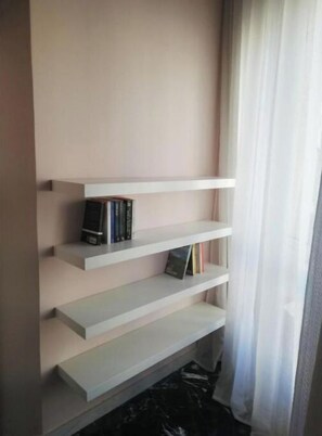 A minimalist bookshelf with three shelves against a plain wall, next to a window with sheer curtains. - Tiber House - Syracuse (Siracusa)