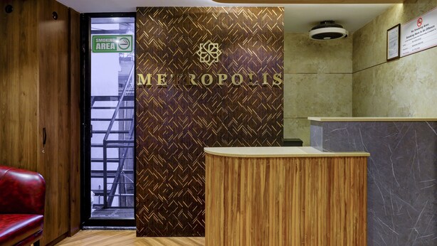 A reception area with a wooden reception desk, a red leather couch, and a patterned wall with the word "METROPOLIS". - Metropolis Business Hotel (Panaji)