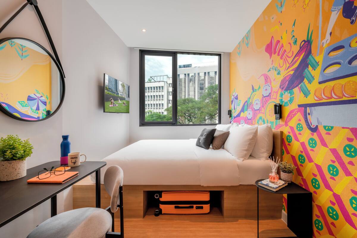 A modern bedroom with a bed, a desk, a chair, a window, and a colorful mural on the wall.