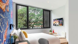 Une chambre d’hôtel moderne dotée d’une grande fenêtre, d’un lit, d’un bureau avec une chaise, d’une télévision et d’une petite table sur laquelle est posée une plante.