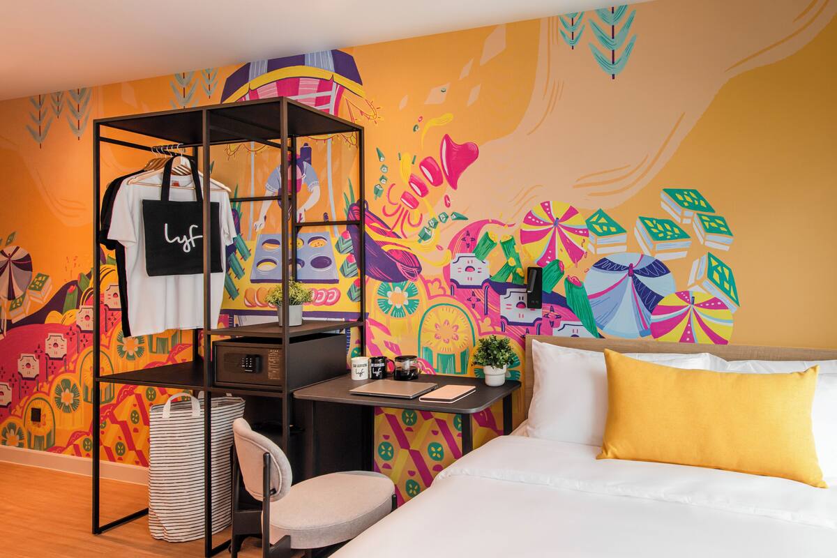 A room with a bed, a desk, a chair, a hanging rack with clothes, and a colorful mural on the wall.