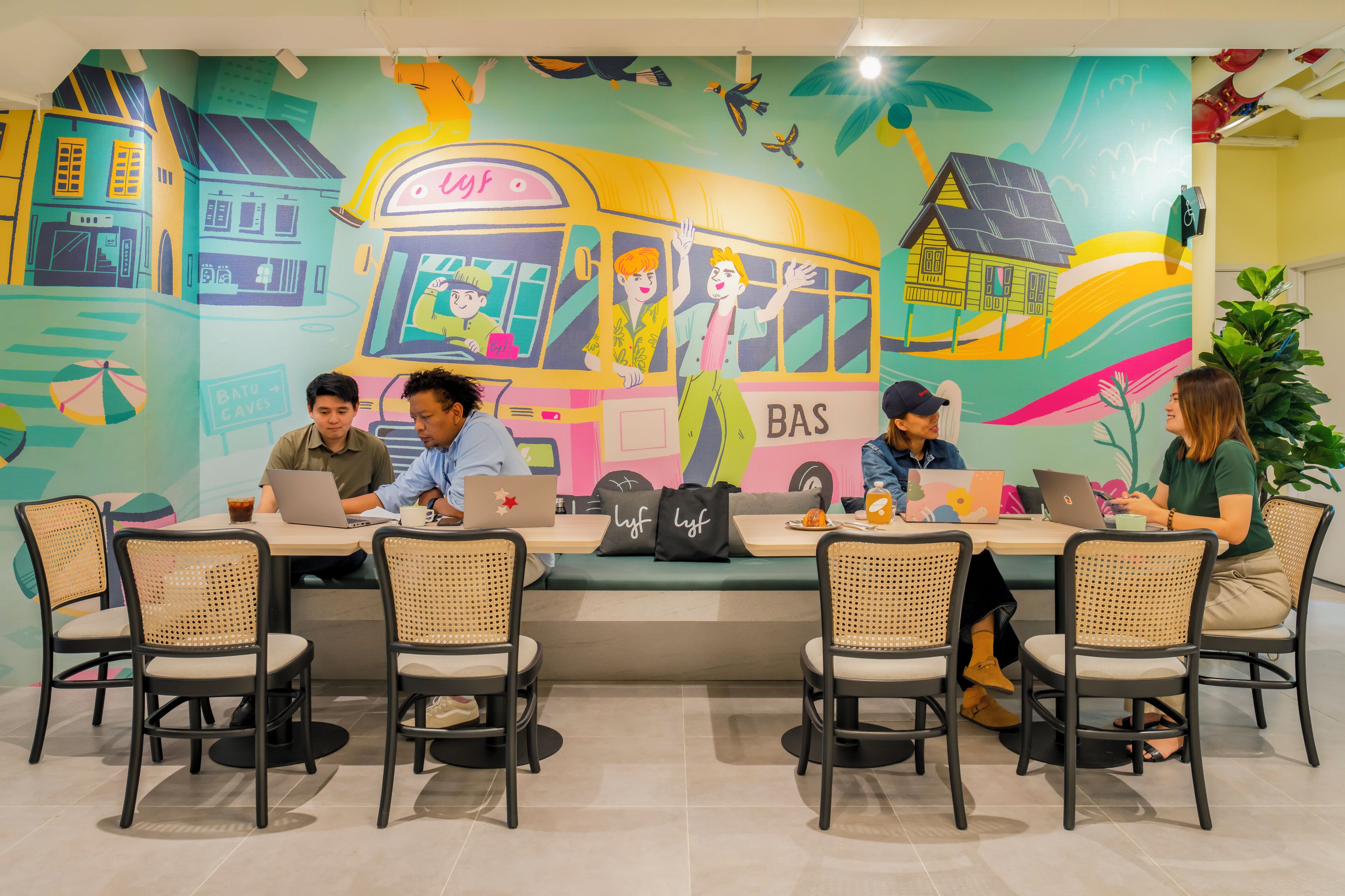 A group of people working at a long table with laptops, surrounded by wicker chairs, in a room with a colorful mural on the wall.