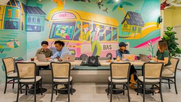 A group of people working at a long table with laptops, surrounded by wicker chairs, in a room with a colorful mural on the wall.
