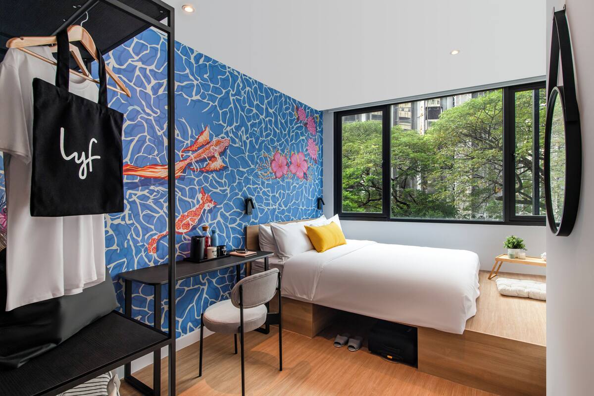 A modern bedroom with a bed, a desk, and a chair. There is a window with a view of trees and buildings.