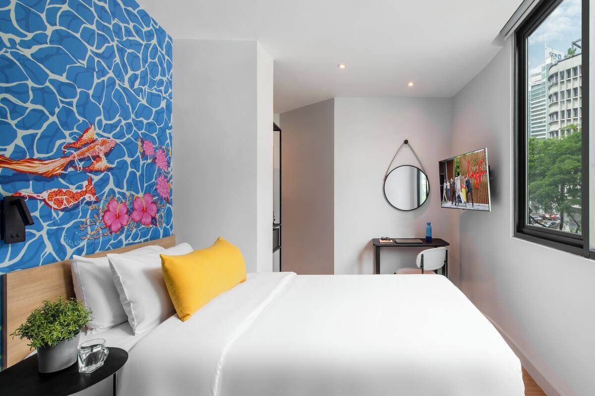 A modern hotel room with a large bed, a small desk, a chair, a wall-mounted TV, and a large window with a city view.