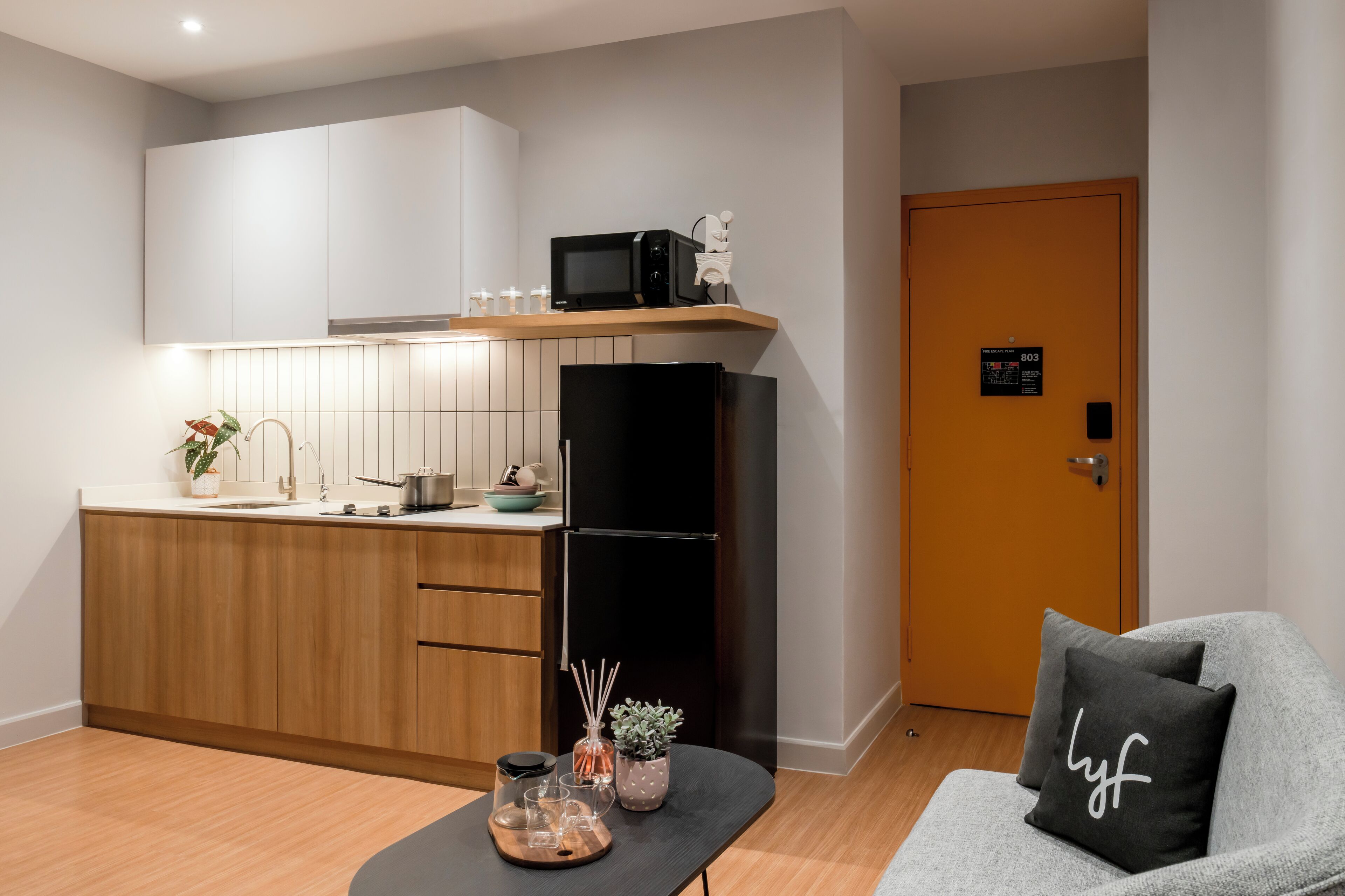 A modern kitchen with wooden cabinets, a black refrigerator, and a microwave on a shelf. There is a door with a sign, a gray sofa with a black pillow, and a small table with a tray and a plant.