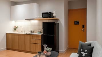 A modern kitchen with wooden cabinets, a black refrigerator, and a microwave on a shelf. There is a door with a sign, a gray sofa with a black pillow, and a small table with a tray and a plant.