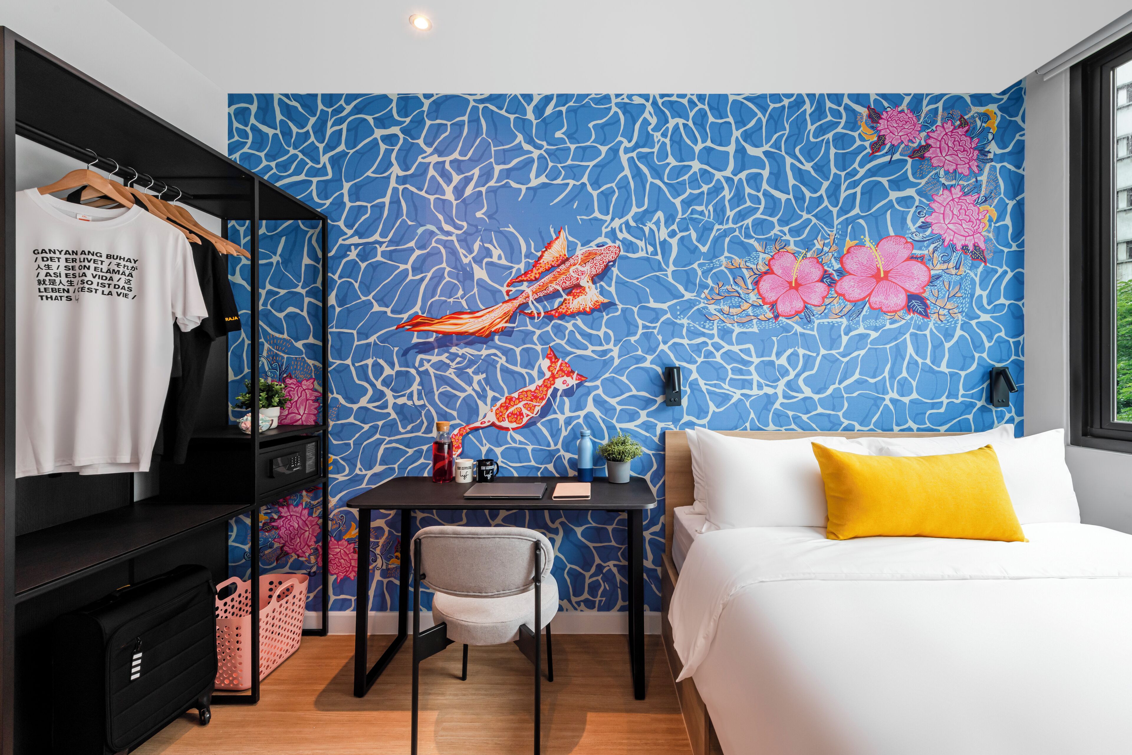 Ett hotellrum med en säng, ett skrivbord, en stol, en garderob och en vägg med en blå, abstrakt design.