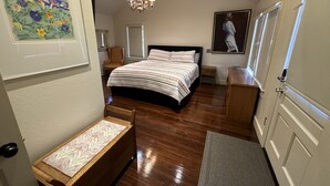 Een slaapkamer met een bed, een fauteuil, een bureau, een kleed, een schilderij, een kroonluchter en een deur.