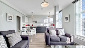 A modern living room with grey sofas, a glass coffee table, and a dining area with white chairs. A kitchen with white cabinets and a dining table is visible in the background. - Roomspace Apartments - Brewers Lane (Richmond)