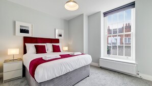 A modern hotel room with a large bed, white bedding, red pillows, a grey headboard, and a large window with a view of buildings. - Roomspace Apartments - Brewers Lane (Richmond)
