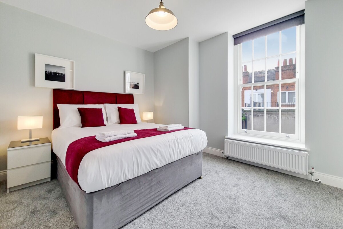 A modern hotel room with a large bed, white bedding, red pillows, a grey headboard, and a large window with a view of buildings.