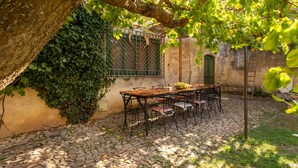 Une cour dallée de pierres, avec une table et des chaises en bois, entourée de verdure et bordée d’un bâtiment à l’aspect rustique.