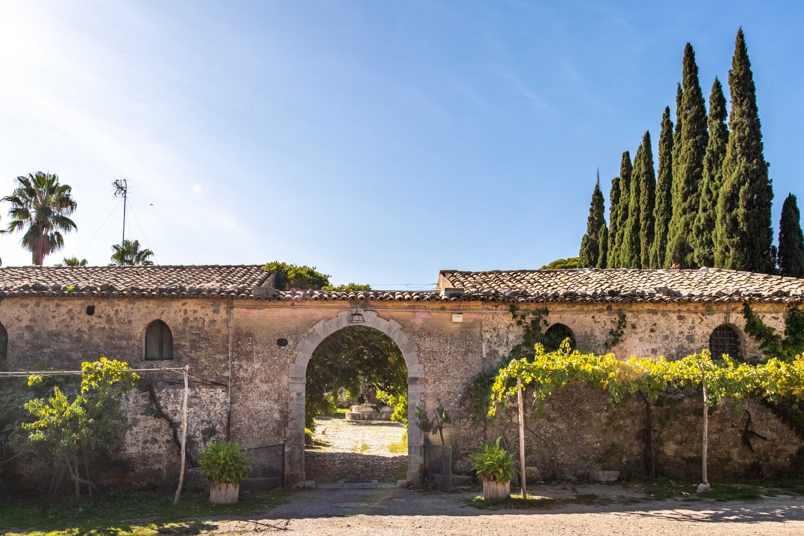 A rustic stone building with an arched entrance, surrounded by greenery and tall cypress trees.
