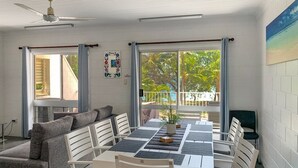Un espace repas comprenant une longue table, des chaises blanches, un canapé gris et un ventilateur de plafond.