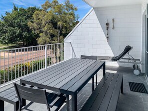 A balcony with a table and chairs, a white wall with a brick pattern, and a view of trees and a road.