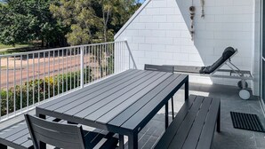 Un balcon avec une table et des chaises, un mur blanc orné d’un motif en briques, et une vue sur des arbres et une route.