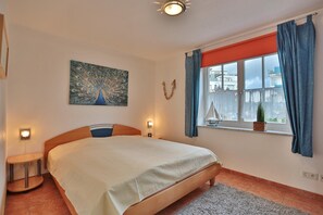 Une chambre à coucher avec un lit, une fenêtre avec des rideaux, un tableau représentant un paon et une petite table avec une lampe.