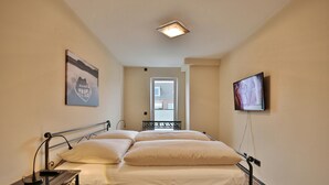 Ein Schlafzimmer mit Bett, Nachttisch, Lampe, Kunstwerk, Fernseher und einem Fenster.