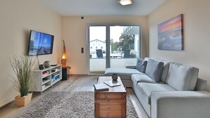Ein modernes Wohnzimmer mit grauem Sofa, einem Flachbildfernseher, einem Holztisch und einem großen Fenster mit Blick nach draußen.