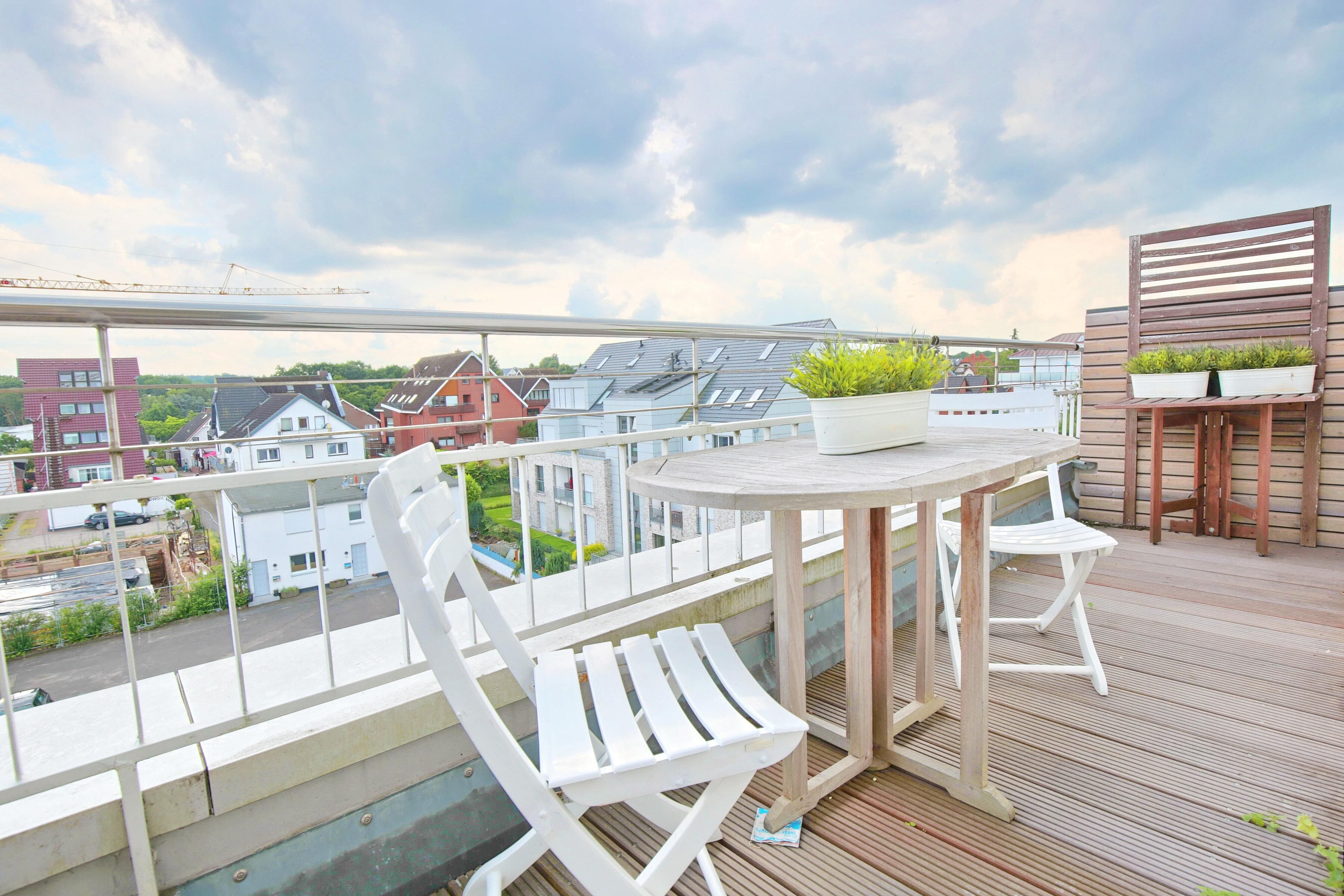 A balcony with a wooden table and white chairs, a wooden bench with potted plants, and a view of buildings and greenery.