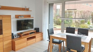 Een moderne woonkamer met een televisie, een houten tv-meubel en een eethoek met een tafel en stoelen.