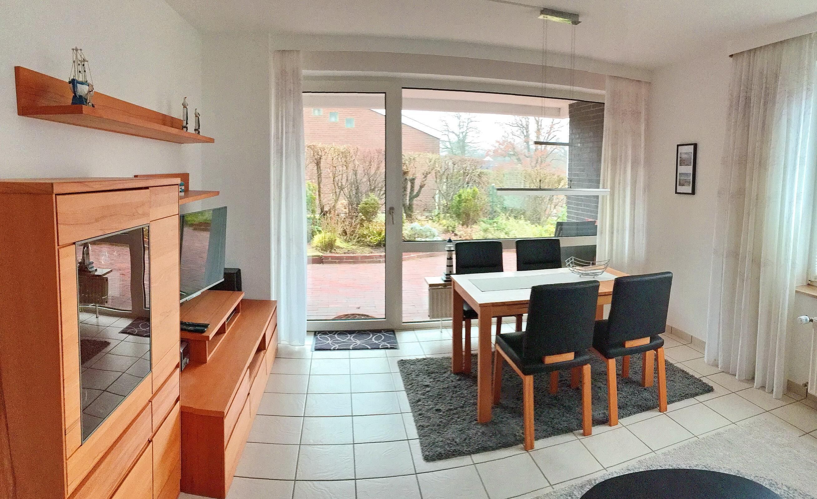 Una sala moderna con televisor, muebles de madera, una mesa de comedor con sillas y un amplio ventanal corredero que da a un jardín.