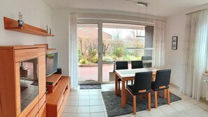 Een moderne woonkamer met een televisie, houten meubels, een eettafel met stoelen en een grote schuifpui naar een tuin.