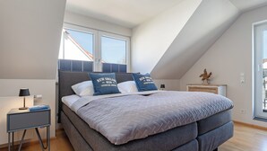 A modern bedroom with a large bed, two pillows with text, a nightstand, and a window with blinds. - Seestern Appartement 3 (Sierksdorf)