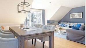 A modern living room with a dining area, featuring a wooden dining table, grey upholstered chairs, a grey sofa, and a large window with a view of the outside. - Seestern Appartement 3 (Sierksdorf)