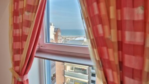 A window with pink curtains, offering a view of a coastal area with buildings and the sea. - Hansa-residenz I Appartement 17 Kumweer (Scharbeutz)